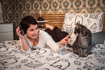 Beautiful woman plays with the cat on the bed. The young woman listens to music on headphones while she is petting her cat.