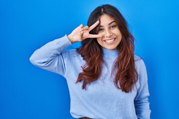 Hispanic young woman standing over blue background doing peace symbol with fingers over face, smiling cheerful showing victory