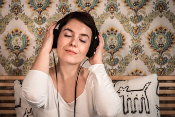 Beautiful woman listens to music in headphones on the bed. The woman has her eyes closed and she is dreaming as she listens to her favorite song about her. - 561596939