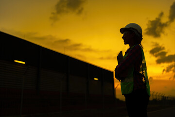 Silhouette of a female engineer smiling. golden sky during sunset standing holding tablet Wear a...