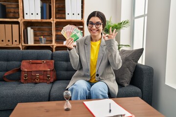 Young hispanic woman working at consultation office holding money doing ok sign with fingers,...