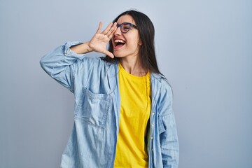 Young hispanic woman standing over blue background shouting and screaming loud to side with hand on mouth. communication concept. - obrazy, fototapety, plakaty