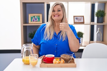 Caucasian plus size woman eating breakfast at home doing happy thumbs up gesture with hand. approving expression looking at the camera showing success.