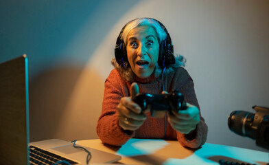 excited elderly woman Gamer in headphones is playing a first-person shooter online video game on her computer. focused on winning in streaming. Cozy night at home.