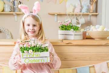 Frohe Ostern. Cute little easter girl laughing with bunny ears on easter day holding basket with text Frohe .Ostern and spring flowers on home kitchen background.