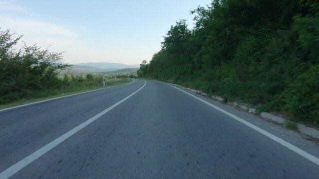 Driving A Car Or Motorcycle Slowly Down On A Winding Mountain Road. Mountain Peaks And Village Are Visible In The Distance. Blue Sky, Summer Time. POV Shot