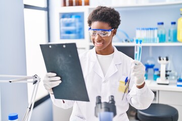 African american woman wearing scientist uniform reading document holding test tubes at laboratory