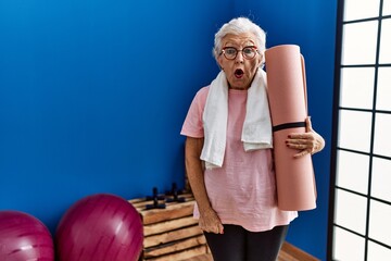 Senior woman with grey hair holding yoga mat scared and amazed with open mouth for surprise,...