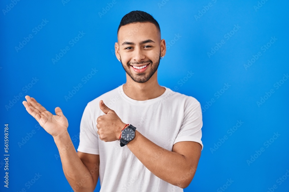 Poster Young hispanic man standing over blue background showing palm hand and doing ok gesture with thumbs up, smiling happy and cheerful