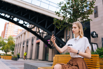 Confident young woman taking selfie using smartphone and holding cup with takeaway coffee in summer day sitting on bench on city street. Cute lady having video call via mobile phone outdoors.