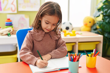 Adorable hispanic girl preschool student sitting on table writing on notebook at kindergarten