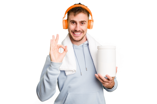 Young fitness man listening to music and holding a protein bottle isolated cut out cheerful and confident showing ok gesture.