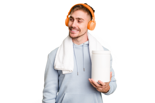 Young fitness man listening to music and holding a protein bottle isolated cut out looks aside smiling, cheerful and pleasant.