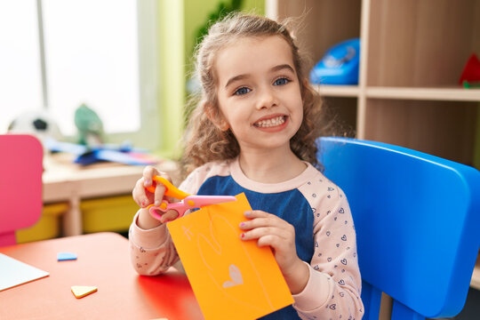 Adorable blonde girl student smiling confident cutting paper at kindergarten