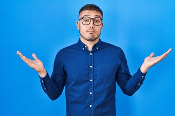 Young hispanic man wearing glasses over blue background clueless and confused expression with arms and hands raised. doubt concept.