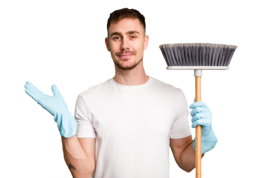 Young man holding a broom to clean his house cut out isolated showing a copy space on a palm and holding another hand on waist.