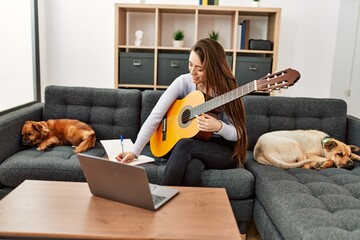 Young hispanic woman having online guitar class sitting on sofa with dogs at home
