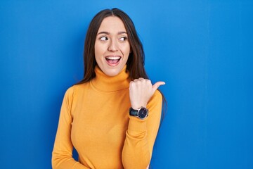 Young brunette woman standing over blue background smiling with happy face looking and pointing to the side with thumb up.