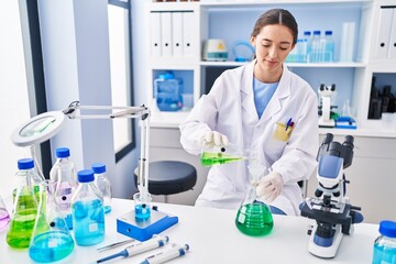 Naklejka premium Young beautiful hispanic woman scientist pouring liquid on test tube at laboratory