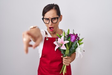 Middle age brunette woman wearing apron working at florist shop holding bouquet pointing displeased and frustrated to the camera, angry and furious with you