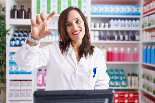 Middle age brunette woman working at pharmacy drugstore smiling and confident gesturing with hand doing small size sign with fingers looking and the camera. measure concept.