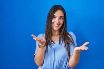 Young brunette woman standing over blue background smiling cheerful offering hands giving assistance and acceptance.