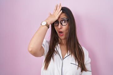 Young brunette woman wearing glasses standing over pink background surprised with hand on head for mistake, remember error. forgot, bad memory concept.