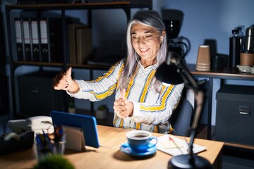 Middle age woman with grey hair working at the office at night approving doing positive gesture with hand, thumbs up smiling and happy for success. winner gesture.