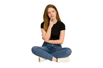 Young redhead woman sitting on the floor cut out isolated looking sideways with doubtful and...
