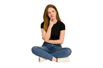 Young redhead woman sitting on the floor cut out isolated looking sideways with doubtful and skeptical expression.
