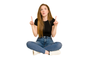 Young redhead woman sitting on the floor cut out isolated pointing upside with opened mouth.