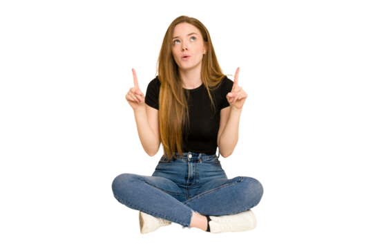 Young redhead woman sitting on the floor cut out isolated pointing upside with opened mouth.