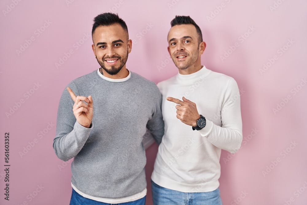 Sticker Homosexual couple standing over pink background with a big smile on face, pointing with hand finger to the side looking at the camera.