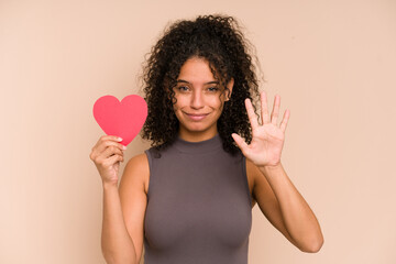 Young african american woman holding a heart for valentines day isolated smiling cheerful showing number five with fingers.