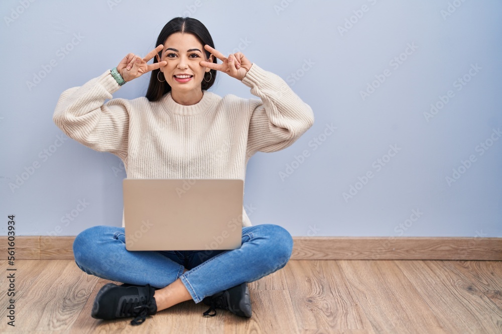 Poster Young woman using laptop sitting on the floor at home doing peace symbol with fingers over face, smiling cheerful showing victory