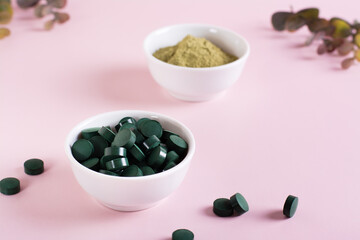 Detox products. Spirulina seaweed in pills and matcha tea powder in bowls on a pink background.