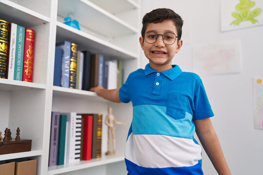 Adorable hispanic boy student smiling confident holding books of shelving at library school