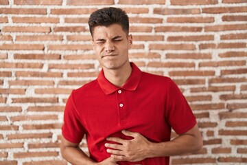 Young hispanic man standing over bricks wall with hand on stomach because nausea, painful disease feeling unwell. ache concept.