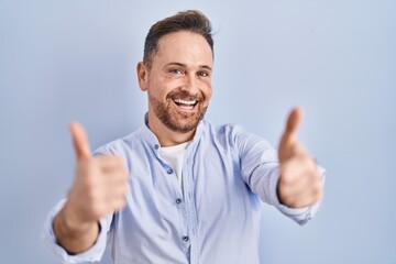 Middle age caucasian man standing over blue background approving doing positive gesture with hand, thumbs up smiling and happy for success. winner gesture.