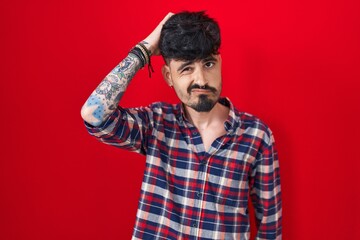 Young hispanic man with beard standing over red background confuse and wondering about question. uncertain with doubt, thinking with hand on head. pensive concept.