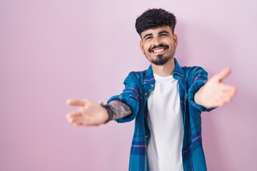 Young hispanic man with beard standing over pink background looking at the camera smiling with open arms for hug. cheerful expression embracing happiness.