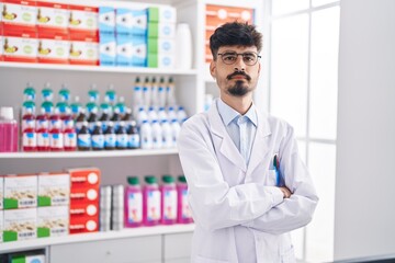 Young hispanic man pharmacist standing with arms crossed gesture at pharmacy