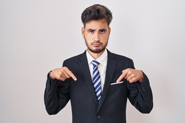 Young hispanic man with tattoos wearing business suit and tie pointing down looking sad and upset, indicating direction with fingers, unhappy and depressed.