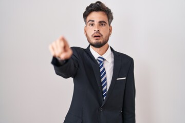 Young hispanic man with tattoos wearing business suit and tie pointing with finger surprised ahead, open mouth amazed expression, something on the front