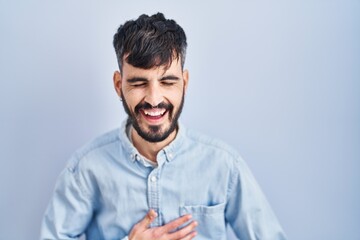 Obraz premium Young hispanic man with beard standing over blue background smiling and laughing hard out loud because funny crazy joke with hands on body.