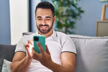 Young hispanic man using smartphone sitting on sofa at home