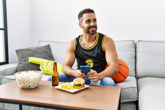 Young Hispanic Man Supporting Basketball Game Using Smartphone At Home