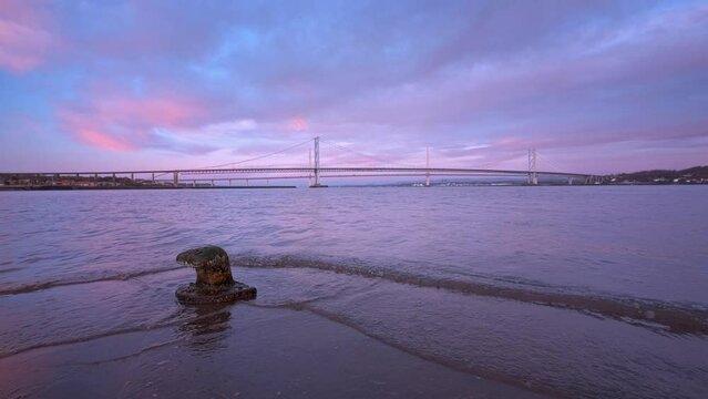 A view of bridge and the sunrise light reflected in the calm sea water and a bollard on the dock closeup. 4k video of a large bridge at sunrise. Forth Road Bridge, Scotland, United Kingdom