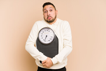 Adult latin man holding a weight scale isolated shrugs shoulders and open eyes confused.