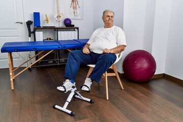 Senior caucasian man at physiotherapy clinic using pedal exerciser looking away to side with smile on face, natural expression. laughing confident.